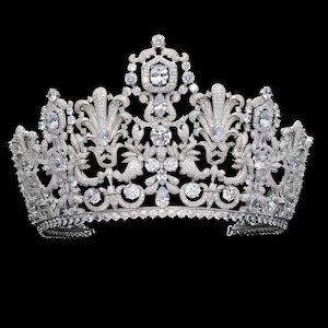 May include: A silver-colored crown with intricate filigree and numerous clear, sparkling gemstones. The crown features a symmetrical design with floral and scroll motifs, set against a black background. The gemstones vary in size and shape, adding to the crown's ornate appearance.