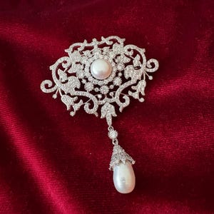 May include: A silver-toned brooch with a large, round pearl center and a dangling pear-shaped pearl. The brooch features an ornate, floral design encrusted with small, sparkling crystals, set against a deep red background.