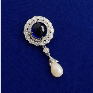 May include: A silver-toned brooch featuring a large, round, dark blue cabochon stone at the centre, surrounded by a halo of clear, sparkling crystals. A single, teardrop-shaped white pearl hangs from the bottom, suspended by a small crystal link.