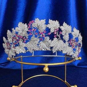Könnte beinhalten: Ein silberfarbenes Diadem mit einer detaillierten Anordnung aus Ahornblättern aus Metall, die mit klaren Kristallen besetzt sind und durch Gruppen kleiner, runder, leuchtend pinker Steine akzentuiert werden. Das Kopfstück hat eine gebogene Basis.
