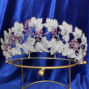 Könnte beinhalten: Eine silberne Tiara mit einem Blatt- und Beerenmuster. Die Tiara ist mit klaren und rosafarbenen Strasssteinen verziert. Die Tiara steht auf einem goldenen Ständer.