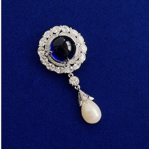 May include: A silver-toned brooch featuring a large, round, dark blue cabochon stone at the center, surrounded by a halo of clear, sparkling crystals. A single, teardrop-shaped white pearl hangs from the bottom, suspended by a small crystal link.