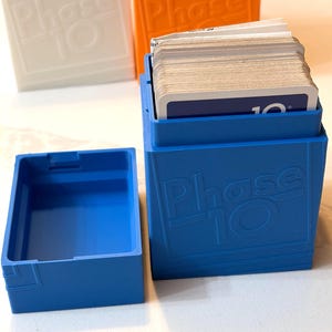 May include: A blue Phase 10 card game box with a lid, next to an open box and two other boxes in white and orange. The boxes have the game name embossed on the front. The open box contains a deck of cards.