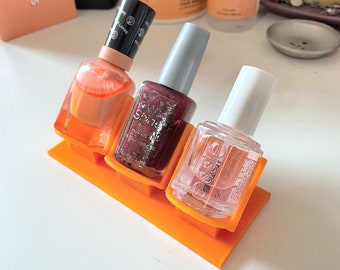 May include: Three clear glass nail polish bottles with orange, burgundy, and pink nail polish. The bottles are displayed on a bright orange plastic stand.