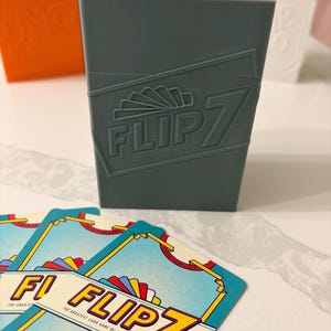 Puede incluir: Una caja de juego de cartas gris con el texto "FLIP 7" y varias cartas con el mismo logo. Las cartas tienen un fondo azul claro con detalles en amarillo y rojo. Se ven otras cajas de juego en el fondo.