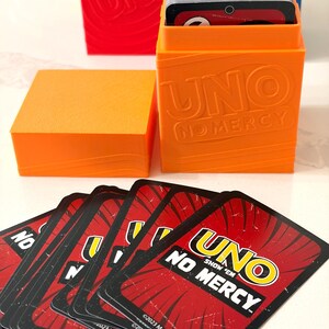 Puede incluir: Un juego de cartas UNO Show 'Em No Mercy naranja. El juego incluye un montón de cartas, una caja de almacenamiento naranja con el logotipo de UNO y una tapa naranja separada. Las cartas tienen un fondo rojo con texto blanco y amarillo.