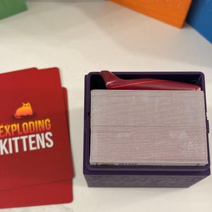 K&ouml;nnte beinhalten: Eine violette Kartenaufbewahrungsbox aus Kunststoff mit einem Kartendeck und einem kleinen roten Haifischflossen-Zubeh&ouml;rteil obenauf. Neben der Box liegen zwei rote Spielkarten mit dem aufgedruckten Text Exploding Kittens und einem Katzensymbol.