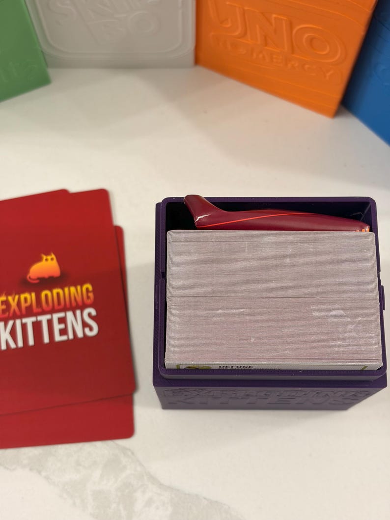 K&ouml;nnte beinhalten: Eine violette Kartenaufbewahrungsbox aus Kunststoff mit einem Kartendeck und einem kleinen roten Haifischflossen-Zubeh&ouml;rteil obenauf. Neben der Box liegen zwei rote Spielkarten mit dem aufgedruckten Text Exploding Kittens und einem Katzensymbol.