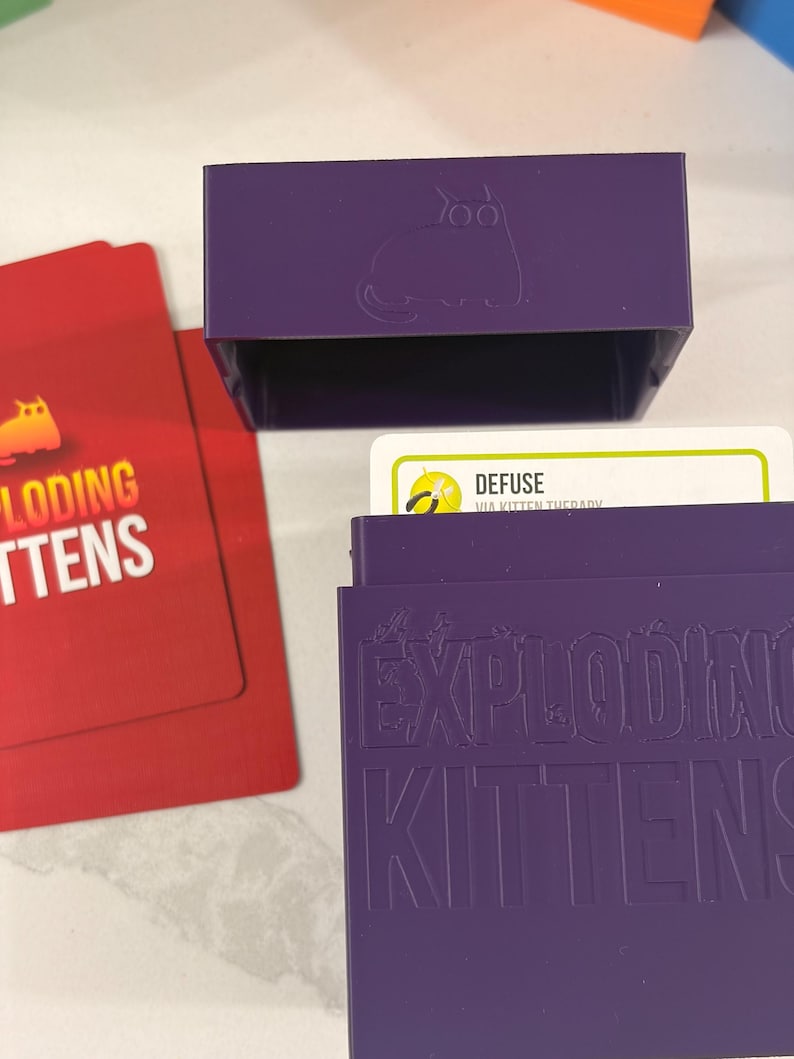 K&ouml;nnte beinhalten: Eine violette Kartenaufbewahrungsbox aus Kunststoff mit dem gepr&auml;gten Text EXPLODING KITTENS und einem Katzensymbol. Daneben liegen rote Spielkarten mit dem Text EXPLODING KITTENS und eine wei&szlig;e Karte mit dem Text DEFUSE VIA KITTEN THERAPY.