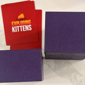 K&ouml;nnte beinhalten: Zwei violette quadratische Boxen mit strukturierter Oberfl&auml;che neben roten Spielkarten. Die Karten zeigen ein gelbes Katzensymbol und den wei&szlig;en Schriftzug EXPLODING KITTENS. Die Boxen dienen als Beh&auml;lter f&uuml;r ein Kartenspiel und sind einfarbig.
