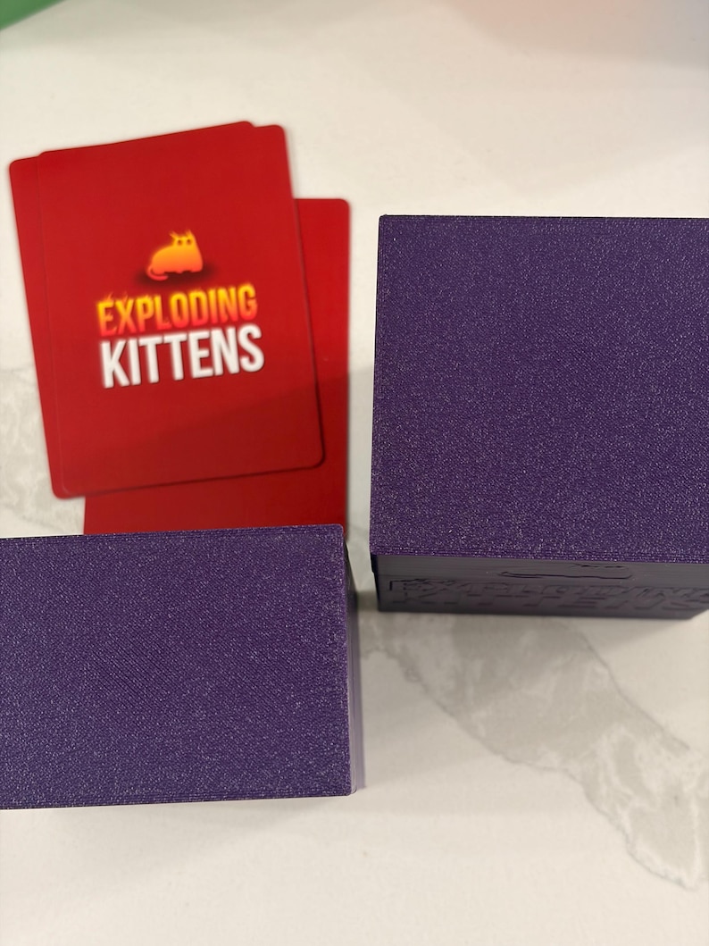 K&ouml;nnte beinhalten: Zwei violette quadratische Boxen mit strukturierter Oberfl&auml;che neben roten Spielkarten. Die Karten zeigen ein gelbes Katzensymbol und den wei&szlig;en Schriftzug EXPLODING KITTENS. Die Boxen dienen als Beh&auml;lter f&uuml;r ein Kartenspiel und sind einfarbig.