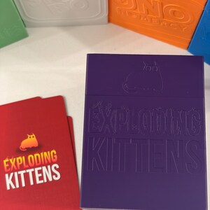 K&ouml;nnte beinhalten: Eine violette, rechteckige Kartenaufbewahrungsbox mit dem gepr&auml;gten Text EXPLODING KITTENS und einem Katzensymbol. Daneben liegen zwei rote Spielkarten mit demselben Text und einer gelben Katze. Weitere bunte Spieleboxen sind im Hintergrund zu sehen.