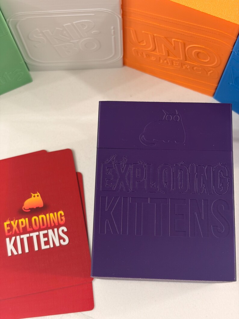 K&ouml;nnte beinhalten: Eine violette, rechteckige Kartenaufbewahrungsbox mit dem gepr&auml;gten Text EXPLODING KITTENS und einem Katzensymbol. Daneben liegen zwei rote Spielkarten mit demselben Text und einer gelben Katze. Weitere bunte Spieleboxen sind im Hintergrund zu sehen.