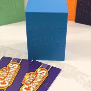 Könnte beinhalten: Eine hellblaue, rechteckige Aufbewahrungsbox für Kartenspiele mit einem geprägten Kronenlogo und dem Schriftzug FIVE CROWNS, begleitet von zwei violett-gelben Five Crowns Spielkarten mit dem Text THE GAME ISN'T OVER 'TIL THE KINGS GO WILD.