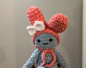 May include: A handmade plush doll with a blue body and a coral-colored hat and scarf. The doll has large, fluffy ears and a white bow. The doll is made of soft, textured yarn and has a smiling face.