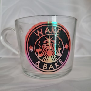 May include: A clear glass mug with a black and pink sticker that says "WAKE & BAKE" with a stylised image of a woman with a marijuana leaf in her hair.