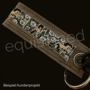 May include: A brown leather keychain with a green and white patterned fabric insert featuring horses and flowers. The keychain has a silver key ring.