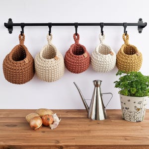 May include: Five hanging crochet baskets in shades of brown, cream, and terracotta, suspended from a black metal rod. The baskets are round with a loop for hanging. A stainless steel watering can and a potted herb plant are also visible.