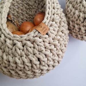 May include: Close-up of two beige crochet baskets, one filled with brown onions. The baskets have a thick, textured weave and a sturdy handle. A small leather tag on the basket reads "Cuddly Island".