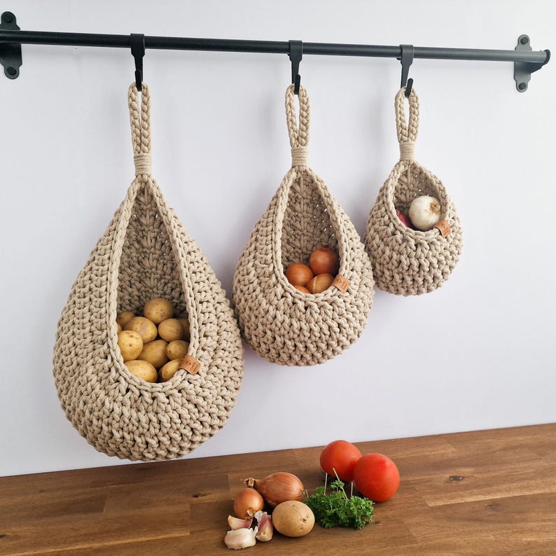 May include: Three beige, teardrop-shaped, crocheted hanging baskets suspended from a black metal rod. The baskets are filled with potatoes, onions, and a garlic bulb. Fresh tomatoes, onions, garlic, and herbs are on the wooden surface below.