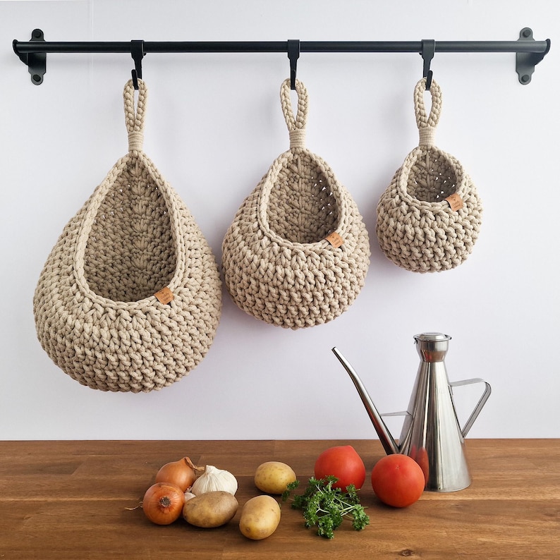 May include: Three beige, crocheted hanging baskets of varying sizes suspended from a black metal rod. The baskets have a teardrop shape with a loop at the top for hanging. A stainless steel oil dispenser and fresh produce, including tomatoes, onions, and potatoes, are on the wooden surface below.