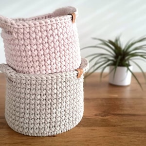 Op de afbeelding: Twee gehaakte manden, een roze en een beige, op elkaar gestapeld. De manden hebben leren handvatten.