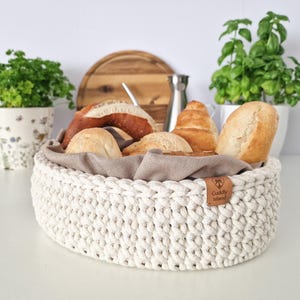 Può includere: Un cestino per il pane bianco, lavorato all'uncinetto, pieno di vari tipi di pane. Il cestino ha un rivestimento in tessuto marrone e un'etichetta in pelle con la scritta "Cuddly Island". Il cestino è su una superficie bianca.
