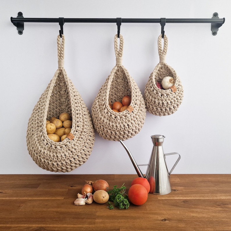 May include: Three beige crochet hanging baskets of varying sizes, suspended from a black metal rod. The baskets are filled with potatoes, onions, and garlic. A metal oil dispenser, tomatoes, and herbs sit on a wooden surface.