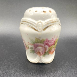 May include: White porcelain salt shaker with a floral design featuring pink and yellow roses. The shaker has a gold trim around the top and a small hole in the top for pouring.