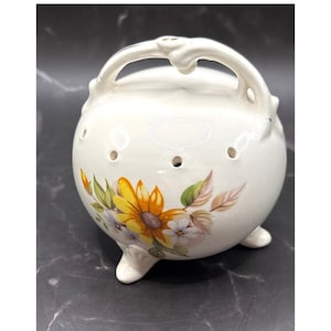 May include: A white ceramic pot with a handle and three small feet. The pot is decorated with a floral design featuring yellow sunflowers and other flowers. The pot has several small holes around the body.