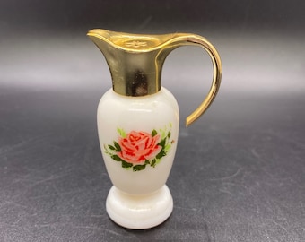 Puede incluir: Una botella de perfume de vidrio blanco con forma de jarra con tapa y asa doradas. La botella tiene una rosa rosa con hojas verdes pintadas en la parte delantera.