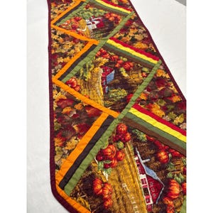 Puede incluir: Un camino de mesa de tela rectangular con un diseño de patchwork de escenas otoñales. El patrón incluye graneros rojos, calabazas naranjas y hojas caídas en tonos marrones, amarillos y verdes, bordeado por un ribete de tela color granate.