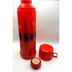 May include: A bright red cylindrical thermos featuring a repeating black diamond pattern. The set includes a matching red screw-on cap with a handle and a separate red stopper with a textured beige circular top, all displayed against a white background.