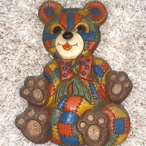 May include: A colorful patchwork teddy bear figurine with a bow tie. The bear is made of various colors, including red, blue, yellow, and green. The bear's paws and face are a light brown color.