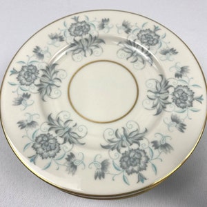 Könnte beinhalten: Ein weißer Keramikteller mit goldfarbenem Rand und einem floralen Design in Grautönen und Blau. Der Teller hat ein zartes Blumenmuster mit einem zentralen Kreis.