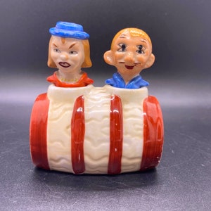 Puede incluir: Un juego vintage de salero y pimentero con una pareja de dibujos animados con rasgos exagerados. La pareja está sentada en un barril de rayas rojas y blancas.