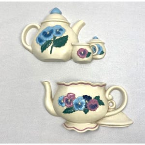 Pode incluir: Conjunto de bule e chávena de cerâmica creme com desenhos florais. O bule apresenta amores-perfeitos azuis, enquanto a chávena tem flores roxas e rosas. O conjunto inclui um açucareiro com uma flor azul.