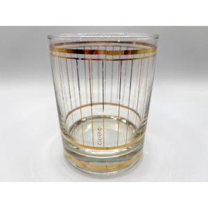 May include: A clear glass tumbler with gold horizontal bands and vertical stripes. The glass has a cylindrical shape and the word "CULVER" is printed on the side. This vintage glassware is suitable for serving beverages.