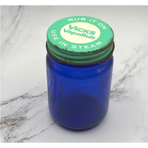 Könnte beinhalten: Ein kobaltblaues Glas mit einem grün-weißen Deckel. Auf dem Deckel steht "Vicks VapoRub" und die Anweisungen "Rub it on, Use in steam". Es handelt sich wahrscheinlich um ein altes Medikamentenglas.