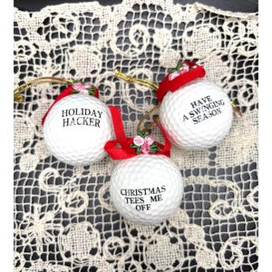 Puede incluir: Tres adornos navideños con forma de pelota de golf blanca con frases humorísticas en texto negro: "HOLIDAY HACKER", "HAVE A SWINGING SEASON" y "CHRISTMAS TEES ME OFF". Cada adorno tiene una cinta roja y un cordón dorado.