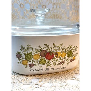 May include: A white oval casserole dish with a clear glass lid. The exterior features a colorful botanical illustration of various vegetables including tomatoes, mushrooms, and artichokes, with the text L'Echalote La Marjolaine printed below.