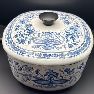 May include: A round, off-white enamel casserole dish with a matching lid featuring a black knob. The exterior of the dish and the lid are decorated with a detailed blue floral pattern, including stylized leaves, blossoms, and intricate border designs.