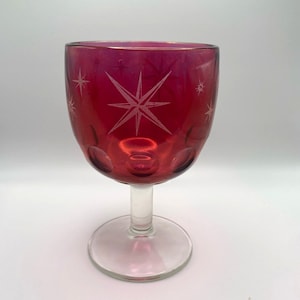 Puede incluir: Una copa de vidrio de color rojo rubí con un tallo y una base transparentes. La copa tiene un patrón de estrella grabado en el vidrio.