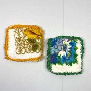 May include: Two crocheted dishcloths with floral designs. One dishcloth has a yellow floral design with a gold crocheted border. The other dishcloth has a blue and green floral design with a green crocheted border.