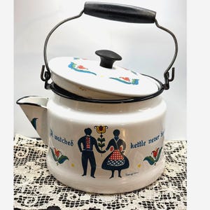 Puede incluir: Hervidor de esmalte blanco con asa y boquilla negras. El hervidor tiene una tapa con un pomo negro y un diseño decorativo. El cuerpo del hervidor tiene un diseño de arte popular con las palabras "A watched kettle never boils".