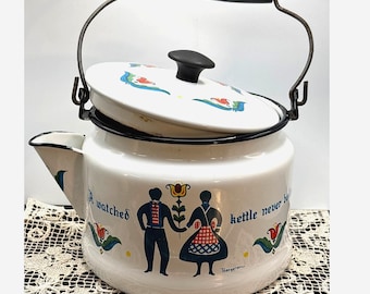 May include: White enamel kettle with a black handle and spout. The kettle features a lid with a black knob and a decorative design. The body of the kettle has a folk art design with the words "A watched kettle never boils".