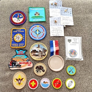 May include: A collection of Boy Scout patches and badges, including a variety of designs and colors. The patches feature images of a compass, a campfire, a wild west scene, and a beaver. The badges include a hiking stick medallion, a first aid badge, and a swimming badge.