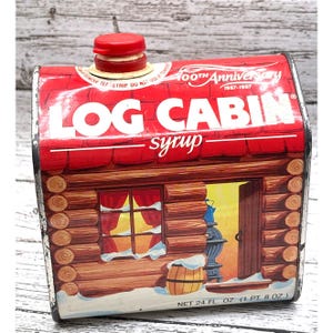 Op de afbeelding: Vintage Log Cabin siroopblik in de vorm van een blokhut. Het rode dak toont "100th Anniversary 1887-1987" en "LOG CABIN syrup". De hut heeft een raam met rode gordijnen, een deur en een besneeuwde grond. Het blik bevat 710 ml.