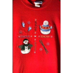 May include: Red sweatshirt with embroidered snowmen and the word "SNOW" repeated multiple times. The snowmen are wearing hats and scarves. One snowman is wearing a blue and white striped sweater.
