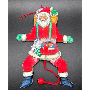 May include: A red and white wooden pull string Santa Claus toy with a green and white stocking, candy cane, and teddy bear in his pockets. He is wearing a green and white striped hat and has a white beard. He is holding a bag of gifts on his back.